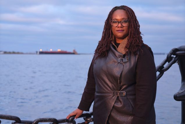 Photo of Deborah Douglas along boston harbor. She wears a black leather jacket, has shoulder-length, crimson locs, and rests her hand on a large middle chain fence. Behind, the harbor at twilight is seen.