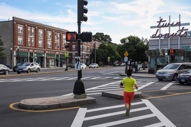 photo of women running through a crosswalk in Allston wearing a neon yellow shirt and orange biker shorts. Twin donuts and various buildings are seen in the background