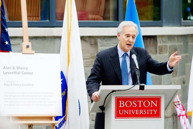 Photo of Alan Leventhal standing at a Boston University podium next to an easel with a poster board that says "Alan & Sherry Leventhal Center." He extends his arm and addresses a crowd during the dedication of the new admissions building, named The Alan and Sherry Leventhal Center on Tuesday, September 16, 2014. Flags are seen behind him.