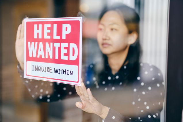 Photo of an Asian woman business owner putting up a help wanted sign in her store window