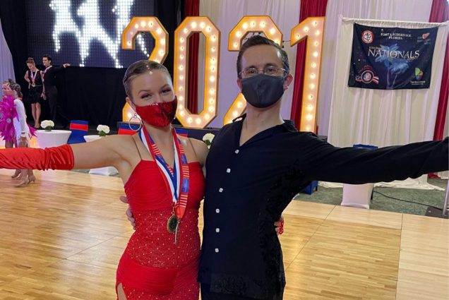 photo of Fiona Brymer had and her partner, Brian Freitas posing after a dance competition. She wears a red dress and he wears black shirt and pants. Both wear blue face masks