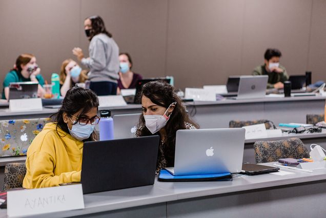 Photo of two students, sitting in a classroom with their laptops open. They wear face masks and one looks at the other's laptop. Students at the back of the classroom talk in a small group.