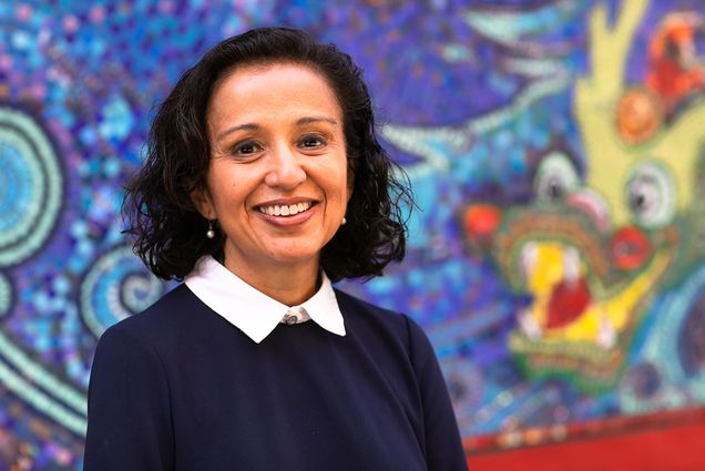 photo of Manjusha Kulkarni, an Indian woman, wearing a blue sweater over a white collared shirt. She stands in front of a bright blue mosaic mural.