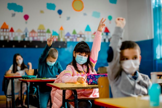 A child wearing a mask raises their hand in a classroom