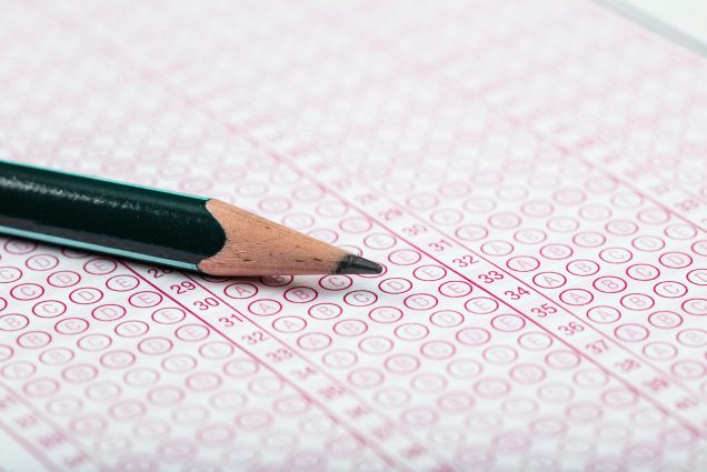 Photo of a green pencil resting on top of a paper with red-ink multiple choice bubbles.