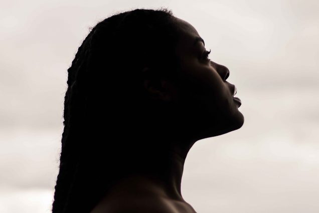 Photo of the side profile of a dark-skinned black woman, bare from the shoulder and with long braids, looks off to the right. The profile is mostly in shadow, though the tones of her skin are noticeable. Background is a hazy white, similar to clouds.