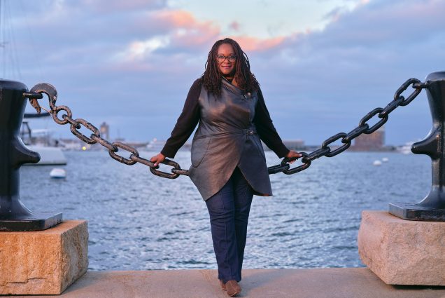 Photo of Deborah Douglas, a dark-skinned Black woman with long dreads wearing stylish glasses and a vegan leather vest, long-sleeved black shirt, and jeans leans back against a large chain fence on a dock. A beautiful purple sunset amidst a partly cloudy sky and large body of water is seen behind her.