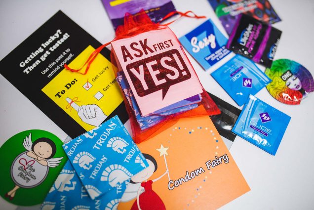 Photo of a variety of different brands of condoms scattered on a white table. Two small postcards included read "Ask first. Yes!" and "Condom Fairy"