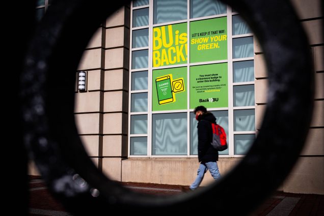 Photo of a student walking past a large BU window on Commonwealth Avenue. Window features a large poster that reads "BU is Back. Let's keep it that way. Show your green"