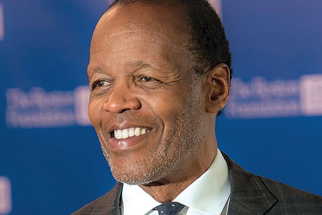 photo of Lee Pelton, a black man with gray stubble wearing a black suit, white shirt, and tie. He looks to the left in front of a darer blue background.