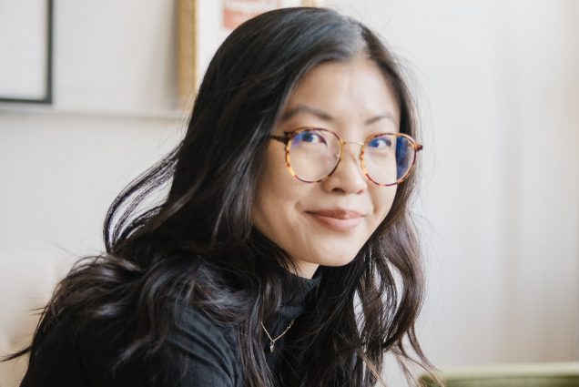 Photo of Weike Wang (GRS’15), a young Asian American woman with shoulder length brown hair and turtle glass. She wears a black blouse and spotted green and yellow skirt and sits with her hands folded as she slightly smiles.