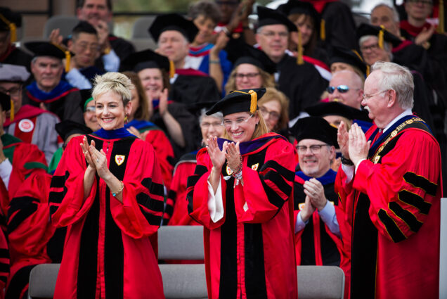 Photo of Ryan Roth Gallo (LAW’99), pictured (left) at the 2018 BU Commencement in full regalia. On the right, Carmen Yulín Cruz Soto (CAS’84) and President Brown are seen. All of them clap, and rows of commencement go-ers in regalia are seen behind them.
