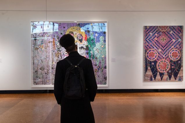 Photo taken from behind Theo Tyson, the Penny Vinik Curator of Fashion Arts at the MFA, regarding the work on display at Life Altering: Selections from a Kansas City Collection, particularly Ebony G. Patterson’s II piece Trees in a Forest. Tyson is a tall, young man of color with his hair pulled up, dressed in black and wearing a black backpack.