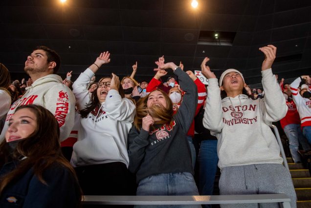 Photo of Mikel Sanchez (CAS’23), from left, Aida Berberian (ENG’23), Maria Paula Fernandez (ENG’23) and Eric Yi (CAS’23) cheer from the dog pound during the Beanpot championship game Feb 14 at TD Garden. They smile, raise their hands in celebrations, and are decked out with BU swag.