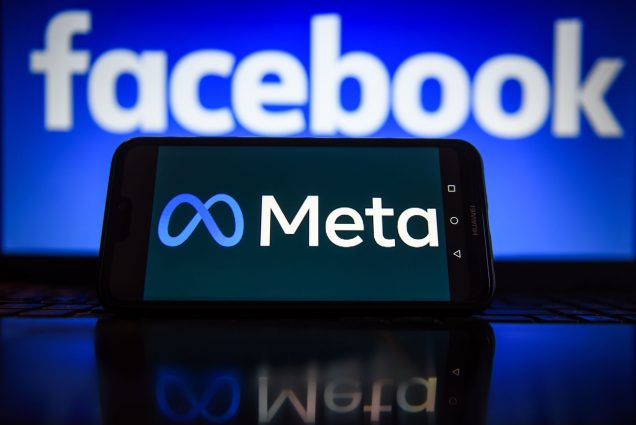 Photo of a phone with the Meta logo on screen. Meta is written in white text to the right of a blue infinity symbol. Behind the phone, a large screen displays the facebook logo.