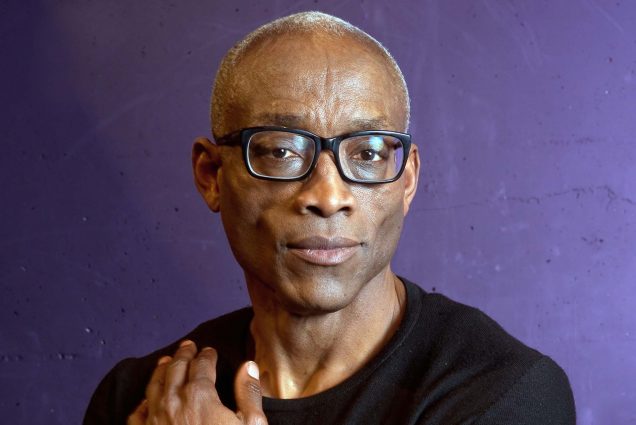 Portrait of Bill T Jones, an older Black man with short white hair, wearing dark glasses, a black sweater. He looks focused and sincere, and touches his right shoulder. He poses in front of a purple background.