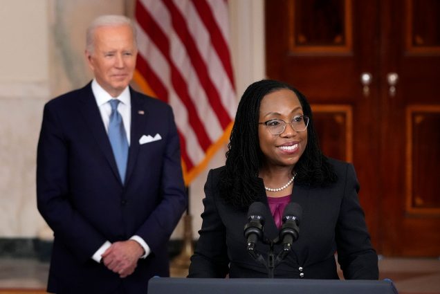 Photo of Ketanji Brown Jackson, a middle aged Black woman in a black blazer, purple blouse, and glasses, smiling as she speaks at a podium during an event in the Cross Hall of the White House February 25, 2022 in Washington, DC. Blurred in the background, president Biden is seen looking on and smiling. Behind him is an American flag.