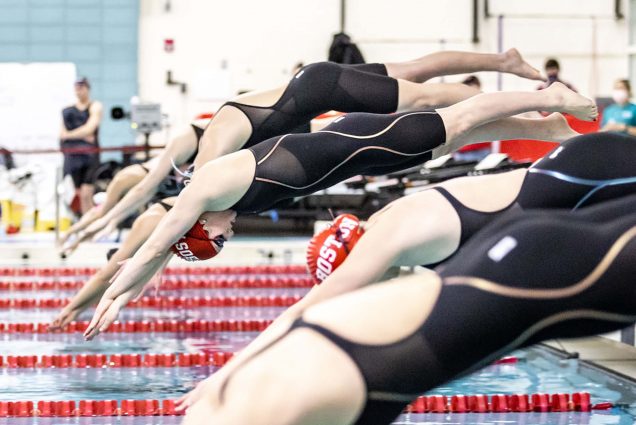 Photo of women in black, full body swim suits and red swim caps, diving into the pool in their given lanes.