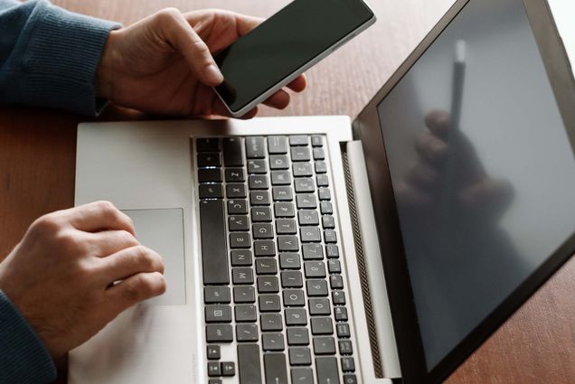 Photo representing dual factor authentication. In the photo, a person holds their iphone in their left hand and uses the trackpad of a mac with the other hand.