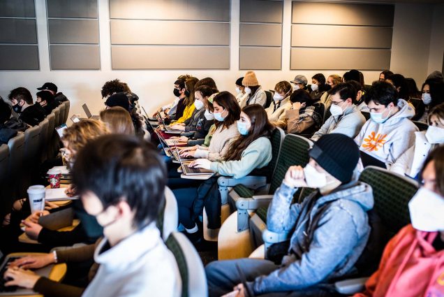 Photo of a class of masked BU students sitting in a lecture hall. They wear a variety of winter coats and outfits as the look to the left towards the front of the room.