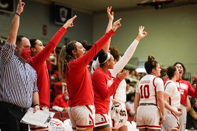Photo of the members of BU Women's Basketball team from the bench cheering. 4 women wearing red long-sleeved shirts hold their arms up and hold their fingers in a dog formation to cheer. A masked ref is seen on the left raising his arm.