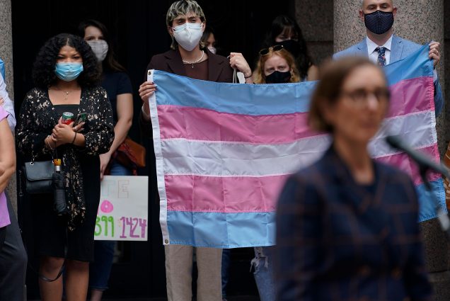 Photo: Parents of transgender children and other supporters of transgender rights gathered in the capital outdoor rotunda to speak about transgender legislation being considered in the Texas House and Texas Senate in Austin, Texas. They hold a large light blue, pink, and white striped flag and wear masks.