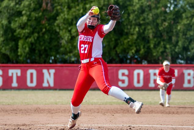 Photo of pitcher Lizzy Avery. A young women in a red BU softball uniform and wearing a helmet is seen mid-pitch in the center of the field on the pitcher's mound.