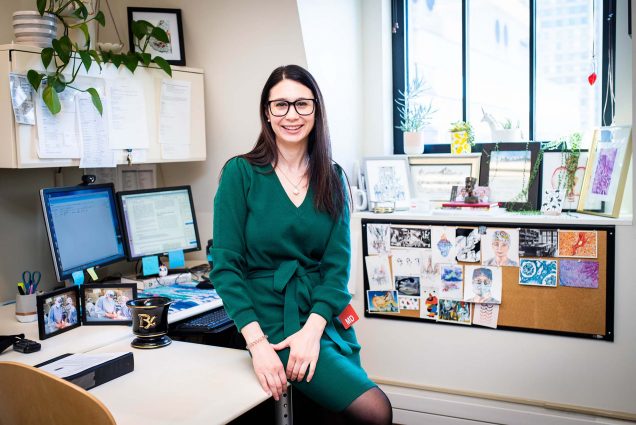 Photo of Andrea Merrill, in a jewel-tone green dress tied in the front, leaning against her desk and smiling. She is light-skinned, has long black hair and wears black glasses. Her red MD badge is seen on her left hip and her office has two monitors and is decorated with lots of photos.