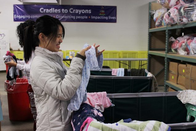 Photo of a young, Asian student in a white puffy picking up a piece of baby clothing from a large bin at a Cradles to Crayon collection center.
