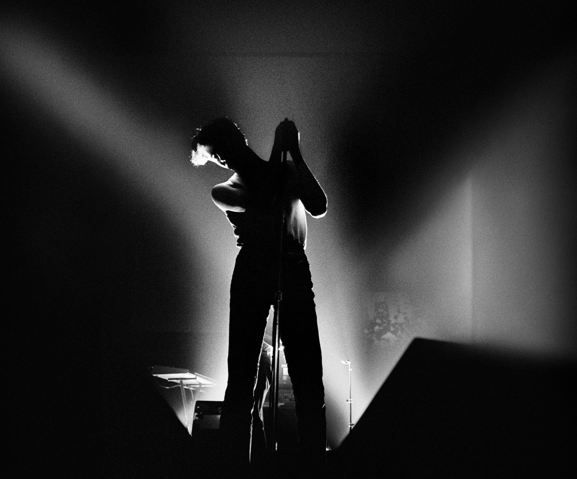 Black and white photo of Susan Ann Sulley of The Human League performing at Spit on Lansdowne Street in Boston. She stands at the mic with bright lights behind her but she’s only seen in silhouette.