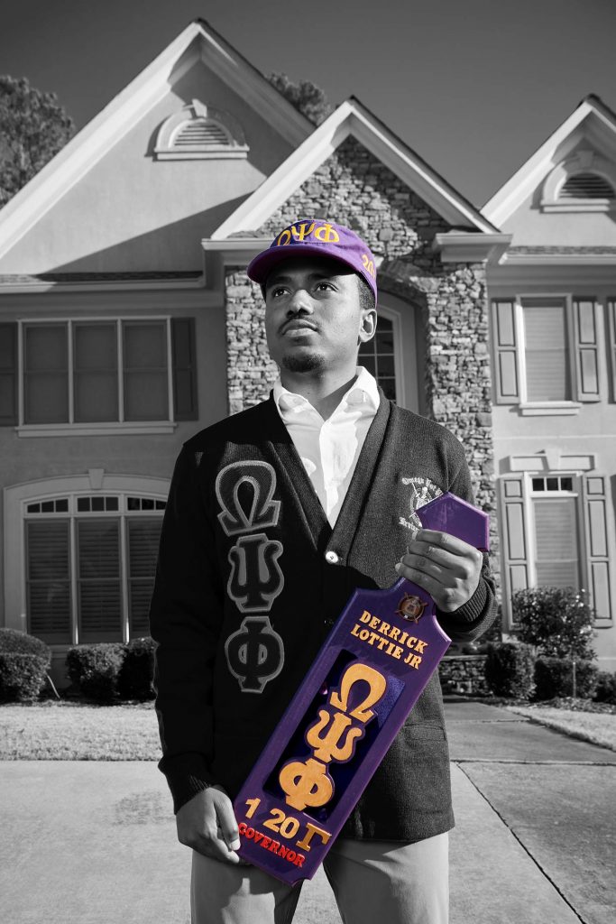 Photo of Derrick Lottie, Jr. (QUESTROM’21) standing on the lawn of a large house. The photo is black and white with the exception of his Brothers United hat and  object that he holds in his hand, both are tinted a bluish purple.