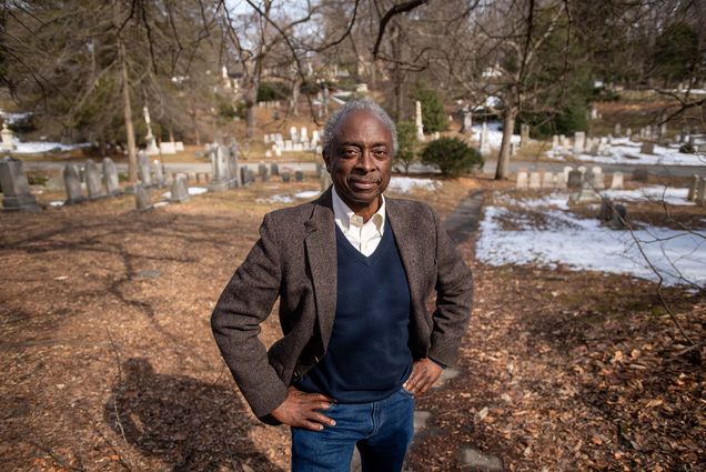 Photo of Roberto Mighty, an older black man with dark grey hair wearing a navy sweater vest over a white collared shirt, with a brown blazer and jeans, stands with hands on hips in front of a cemetery.