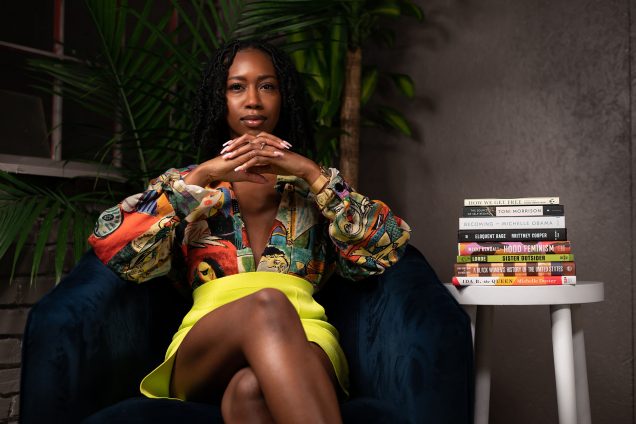 Photo of Lynae Vanee Bogues sitting in a dark blue velvet chair in front of small, potted palm trees with a stack of books by Black authors sitting in a neat pile on a white side table next to her. A young black woman with shoulder length faux locs and a multicolored blouse and bright yellow skirt sits with fingers crossed in front of her chin as she poses seriously for the camera.