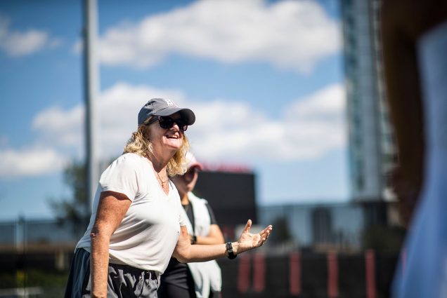 Candid photo of BU women's tennis head coach Lesley Sheehan. She is shown wearing a plain white tee, large black sunglasses, gray cap, and great sweatpants as she smiles and gestures a swing with her hand on a clear, sunny day. A blurry figure is seen behind her.