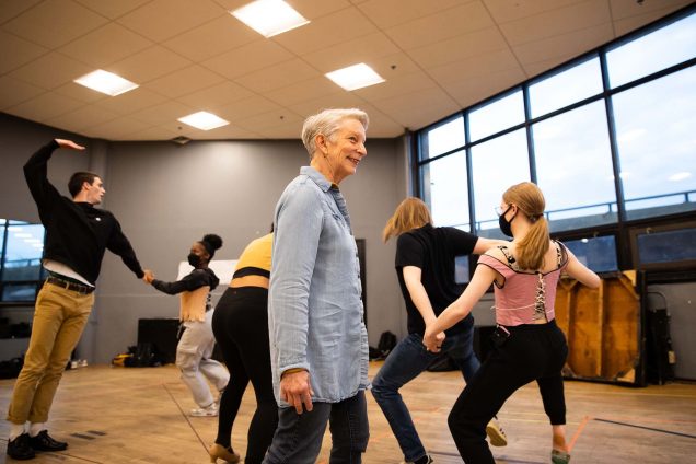 Photo of Judy Braha (CFA’08) at a rehearsal for Shakespeare in Love. Braha wears a long, light blue button-down and black jeans. She's a white woman with short, gray hair. Behind her, in a room with wooden floor, five students are seen rehearsing dance moves.