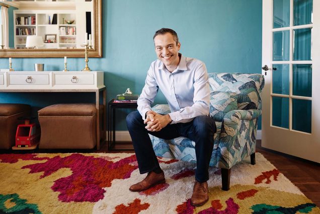 Photo of Nathan Blecharczyk, Airbnb chief Strategy officer, sitting in a chair with a repeating blue and white mountain pattern in a highly stylish room, presumably an Airbnb. Blecharczyk wears a button down, jeans and leather shoes, smiles and clasps his hands, his arms resting on his knees. He is a middle-aged white man with short hair.