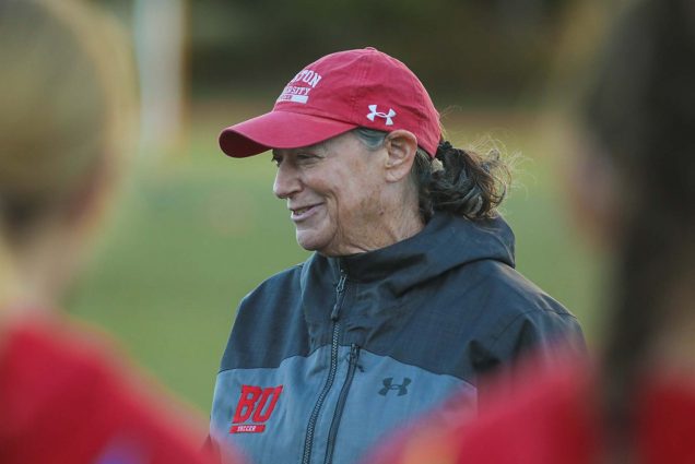Photo of Nancy Feldman, a White middle-aged woman, wearing a red BU hat, and gray BU zip-up. She smiles and looks off to the side. BU women's soccer players are seen blurred in the foreground.