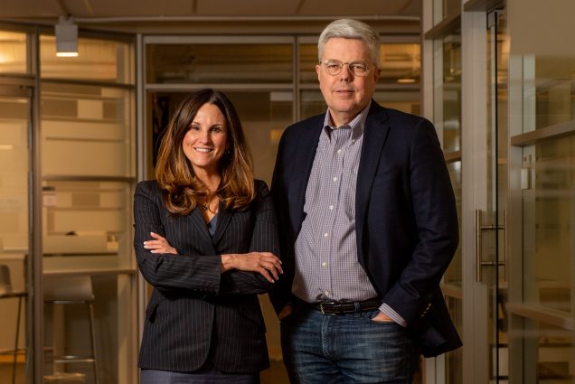 Photo of Kevin Outterson, Executive Director, CARB-X and Erin Duffy, CARB-X's chief of research and development standing side by side at CARB-X. Outterson stands to the left with hands in pocket. He wears a navy blazer, jeans, and a light purple collared shirt. Duffy stands with arms crossed to the left wearing a navy blazer and skirt.