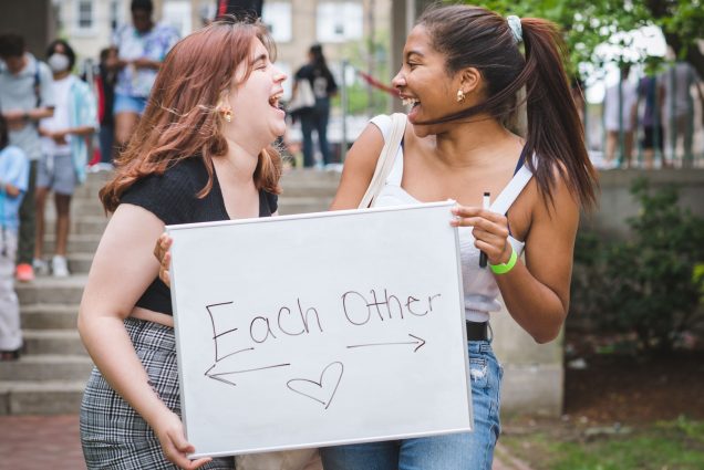 Photo of Natalie Ackerman (COM 22) and Aurouramary Estaba (CAS, Pardee 22) , a young white woman and young black woman. They stand and pose holding a sign that reads "each other" with arrows pointing to each other and a heart emoji. They both look to each other and laugh.