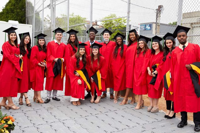 Photo of Graduate Medical Sciences master’s degree candidates before their convocation ceremony. A diverse group of young adults stand in a group wearing red caps and gowns and black caps on a sunny day.