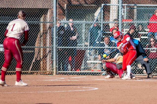 Action photo of Marina Sylvestri. She is caught mid-swing, about to hit a softball thrown by the opposing pitcher wearing a burgundy uniform. Spectators can be seen on the other side of the fence behind homebase, watching on.