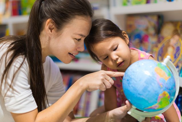 Photo of an Asian parent pointing something out to her child on a small globe in a library bookstore.