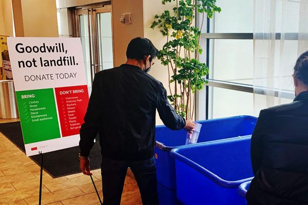Photo of a student putting a water pitcher into a large blue bin near the entrance of a residence hall. Student is dressed in all black and has short hair and sign next to the row of large blue bins reads "Goodwill, Not Landfill" and lists what can and cannot be donated.
