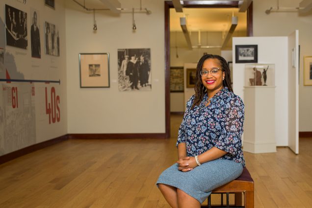 Photo of Melanee Harvey, an assistant professor of art history at Howard University. She is a youngish Black woman, with shoulder-length locs, large cat-eye glasses, who wears a blue floral blouse and a blue knit skirt. She smiles, and clasps her hands on her lap. She sits on a bench in the middle of a gallery. Art hangs on the walls around her.