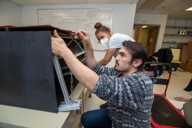 Photo of Tess Ravick (ENG’22), back, and Trevor Melsheimer (ENG’22), front work on their senior design project April 29. The device harvests water from the air. Ravick has light brown skin, wears a t-shirt, a white face mask, and their hair in a bun. Melsheimer, who has light skin and a beard and wears a gray sweater, adjusts a level of the black device on the table in front of them.