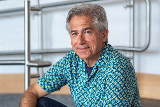 Photo of Armenian-Iranian Albrik Avanessian (MET’22). He is in his 60s, has short gray hair, and has a sincere smile. He wears a blue and white patterned shirt and dark jeans, and sits on a wooden step, resting his arms on his knees.