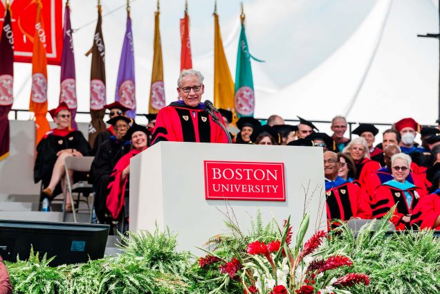 Photo of Bob Woodward during his commencement address. Woodward is an older White man with black-rimmed glasses. Woodward wears a red gown with black. He is seen mid-speaking, at a large white podium with a red "Boston University" sign. A floral arrangement is seen in the foreground; behind him, honorary degree recipients, faculty members, and colored flags are seen blurred. Photo by Melissa Ostrow