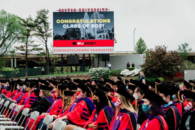 Photo of students sitting on Nickerson field during the 2021 commencement. They wear black graduation caps and red gowns. Behind them, the large jumbotron screen is seen. It reads "Congratulations class of 2021" on screens, and below, a white banner reads "Congratulations graduates!"