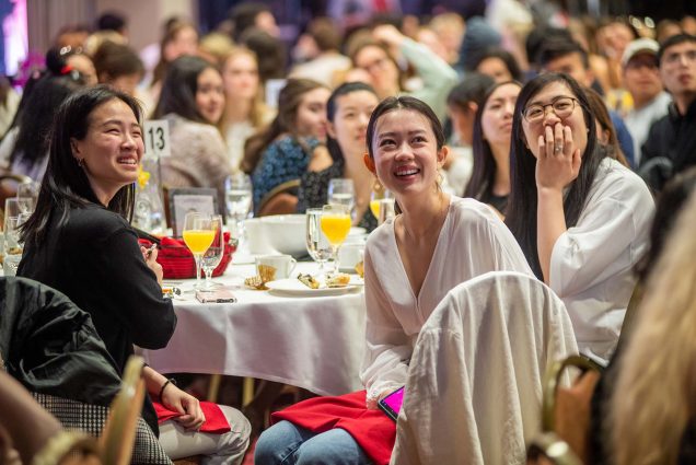Photo of Rebecca Chang (CAS’22), left, June Lin (Questrom’22), middle and Victoria Phang (Questrom’22), right, smiling and laughing as they watching the slideshow during senior class breakfast at the GSU’s Metcalf Ballroom May 5. The three all appear to be AAPI women. Behind them, their table is covered with breakfast food and glasses filled with orange juice.