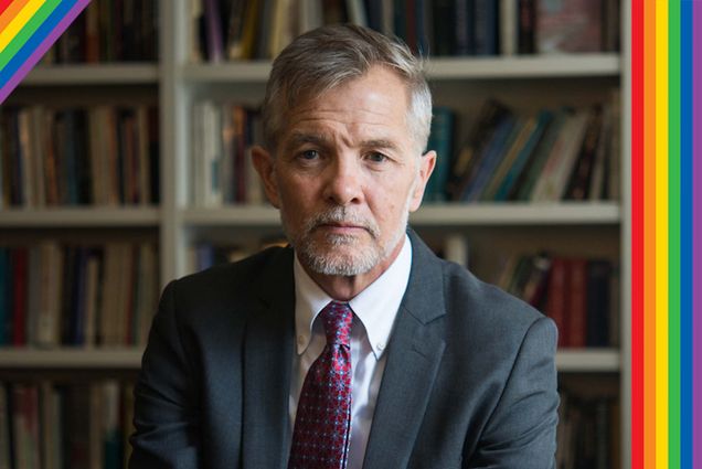 Photo of David Chard, a white man with short gray hair and short gray beard. He wears a blue-gray suit and maroon tie and sits with his hand folded over his knee in what looks like a study or office. At top left and right, rainbows are overlaid.